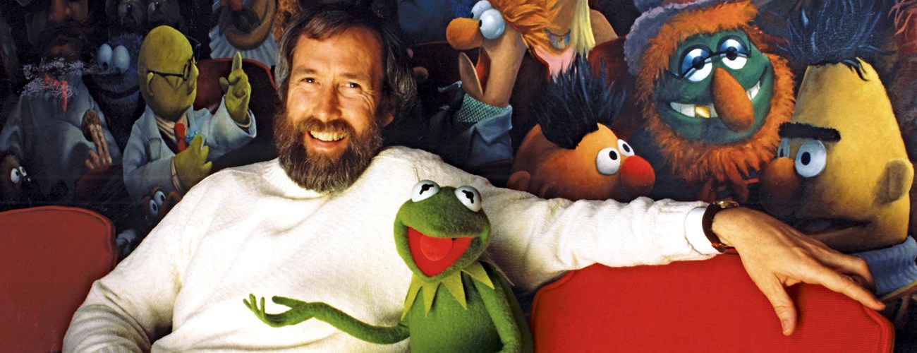 Jim Henson ao lado de Kermit. Os dois estão em frente a um painel com uma pintura de vários personagens.